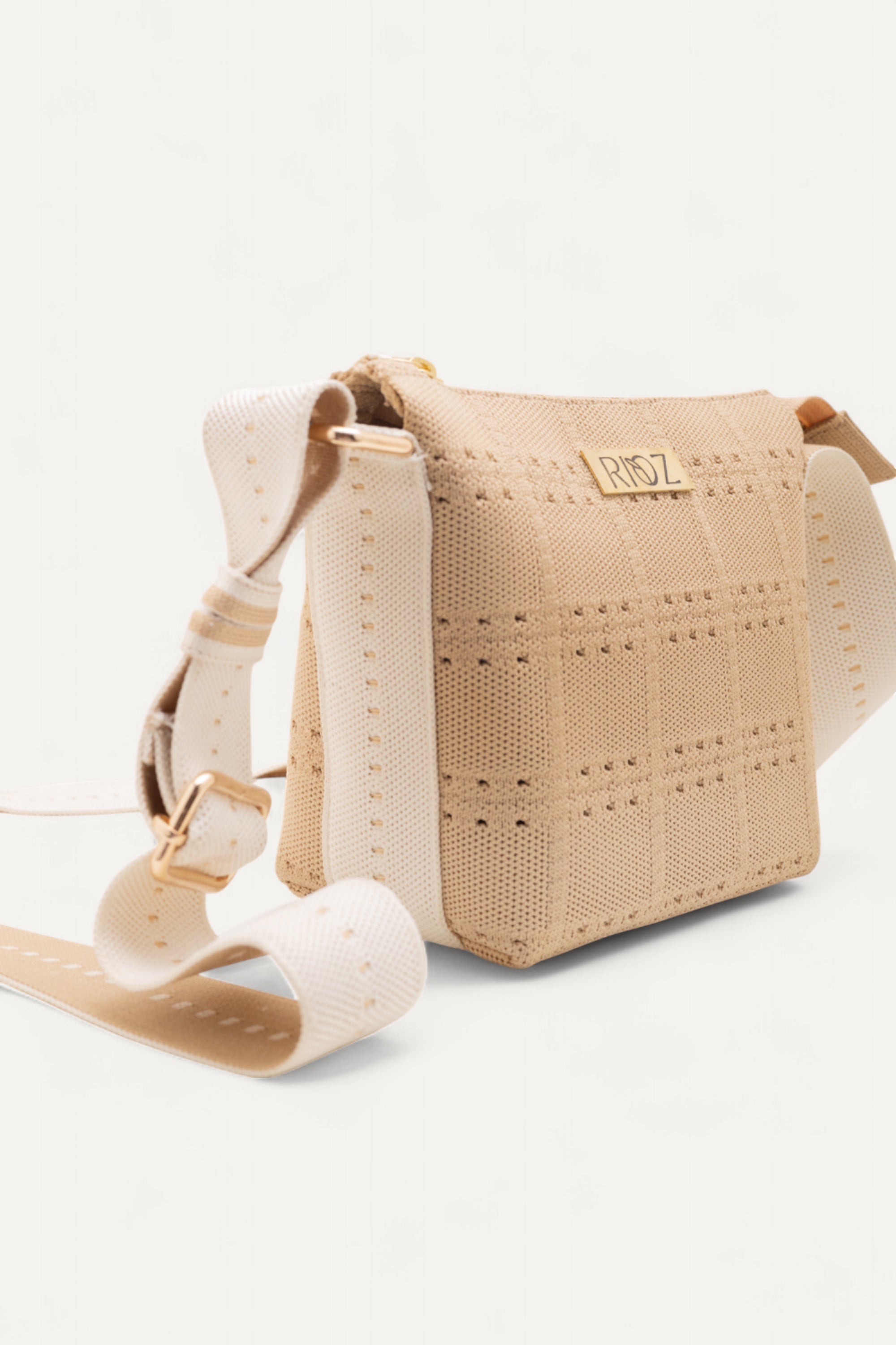 Side view of beige crossbody bag by Rioz with wide white strap; features perforated texture, zip closure and adjustable strap; eco-friendly bag on light gray background.