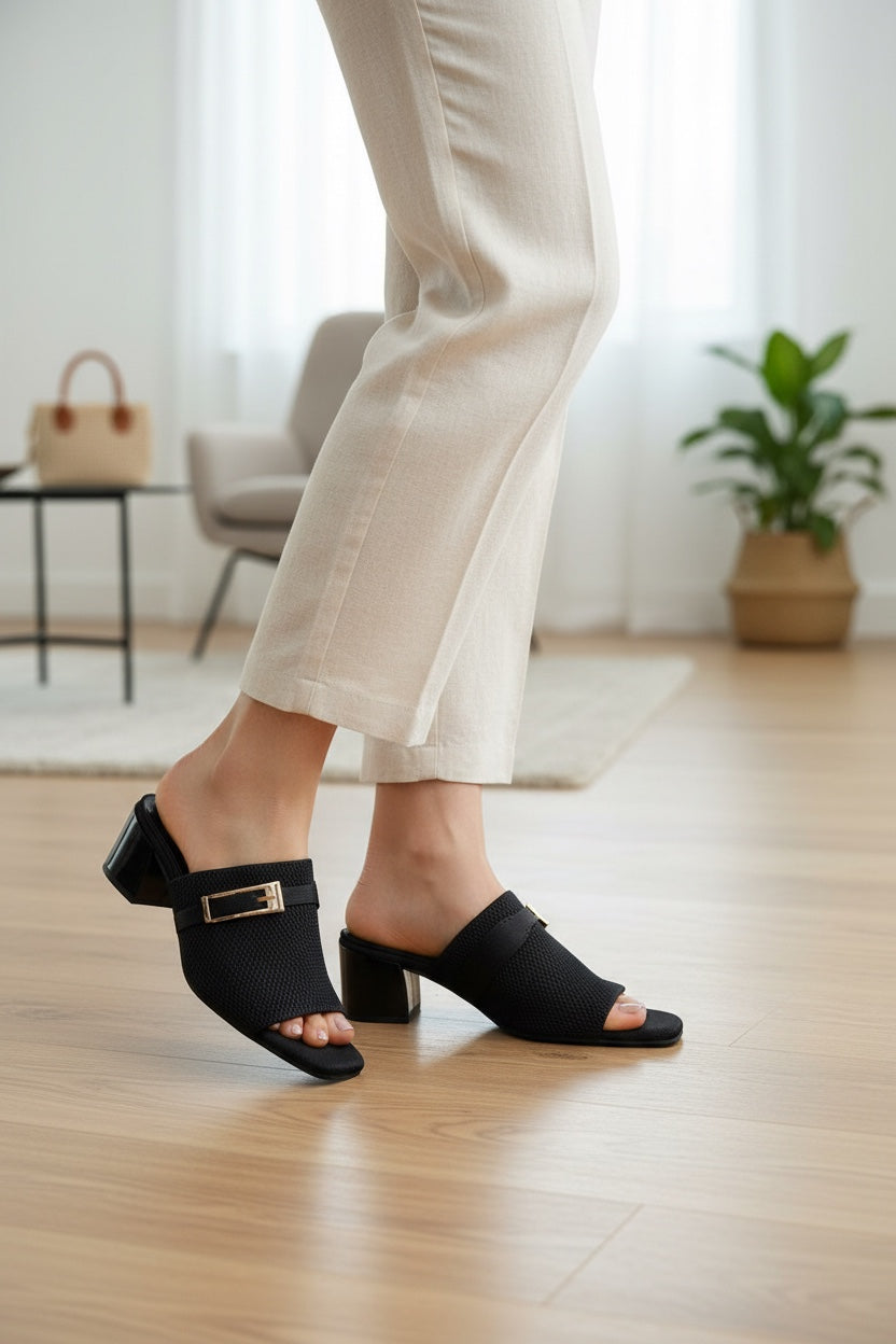 Rioz Nova black knit block heel sandals with a gold-tone buckle detail worn with beige trousers in a bright room.