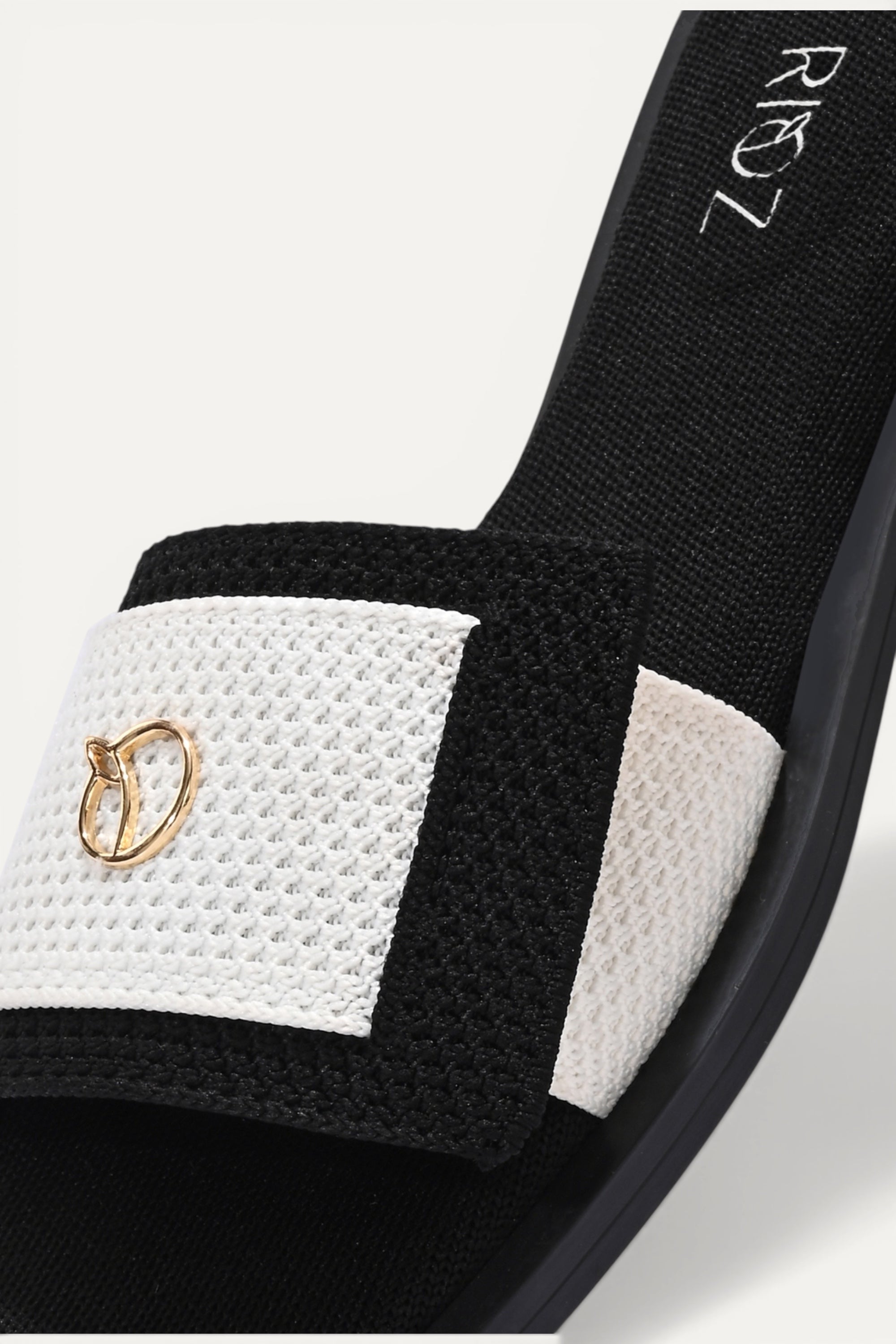 Rioz black and white knit origami slides with gold emblem detail. Asymmetrical fold design for a comfortable, breathable fit.