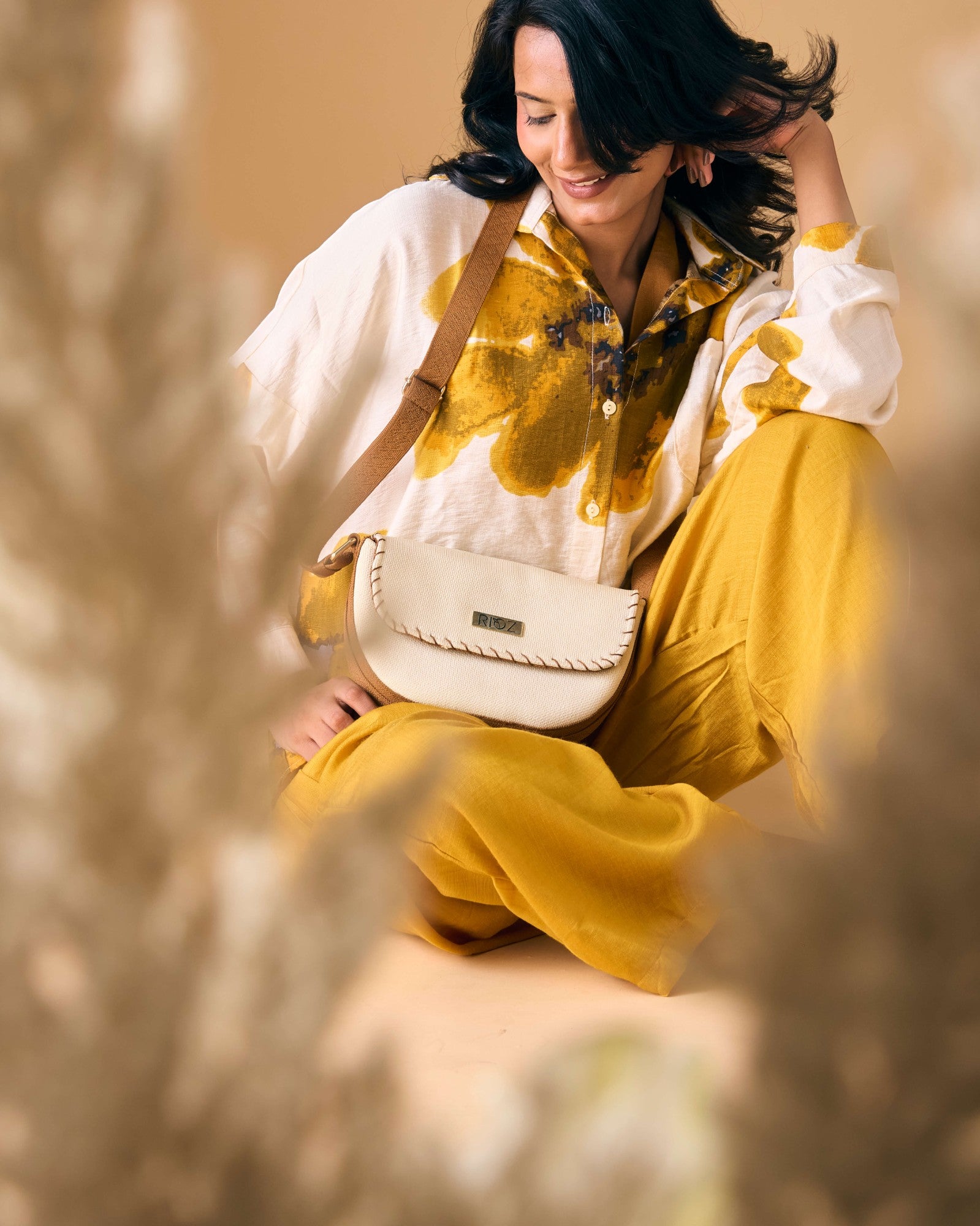 Model sitting cross-legged wearing eco-friendly 9 to 9 Saddle Bag by Rioz across her body with brown strap and ivory bag; sustainable, stylish and compact.