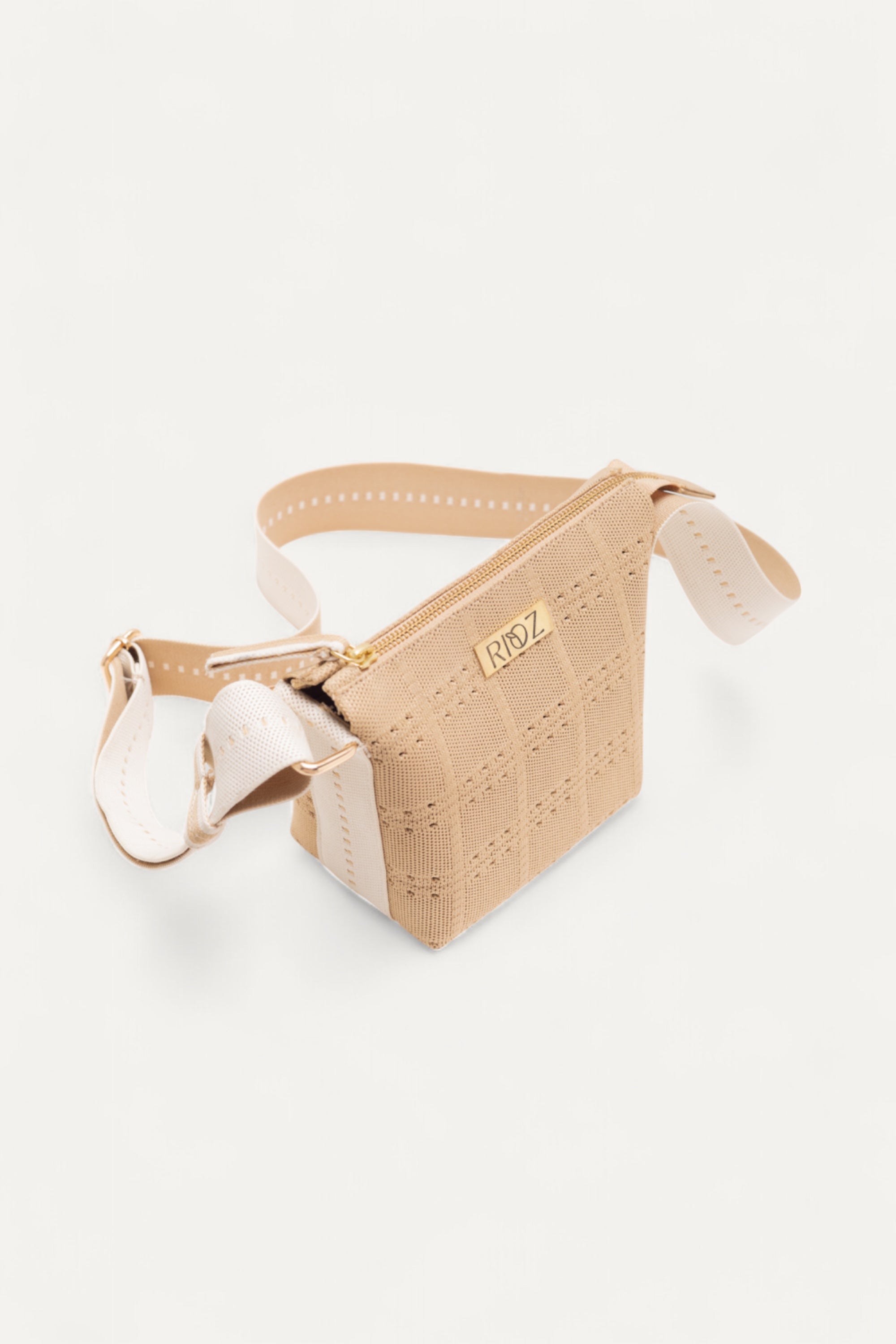Angled view of beige crossbody bag by Rioz with white straps and zip closure; shows perforated woven texture and gold logo; displayed on white background.