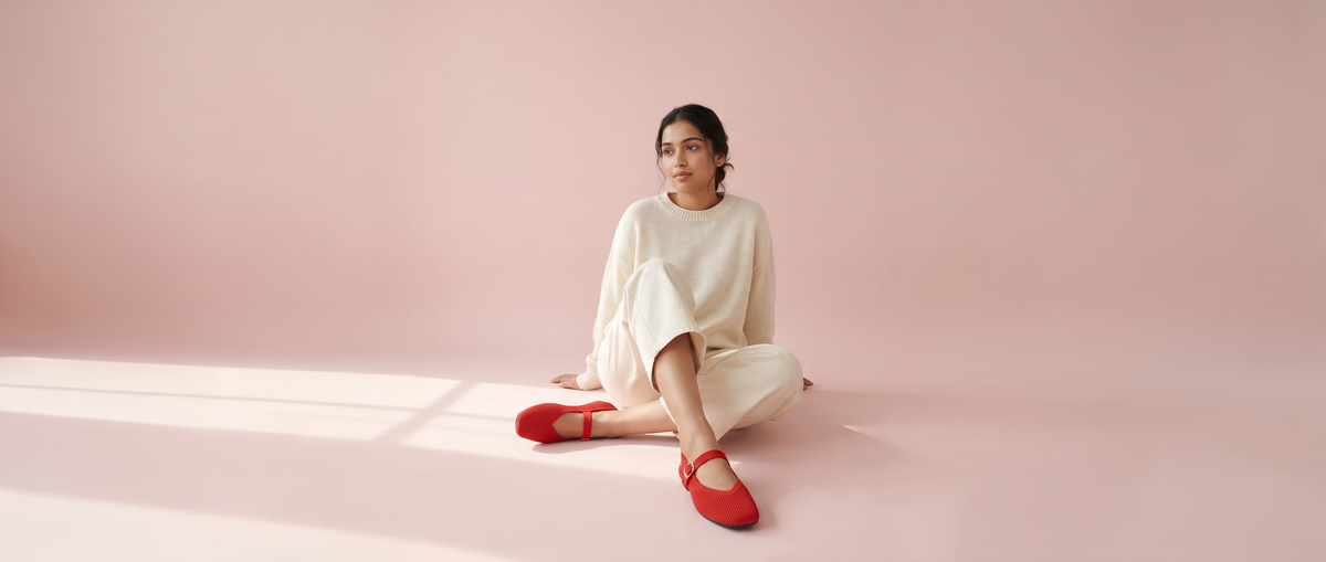 Woman seated on a blush pink studio floor wearing red knitted Rioz Mary Janes