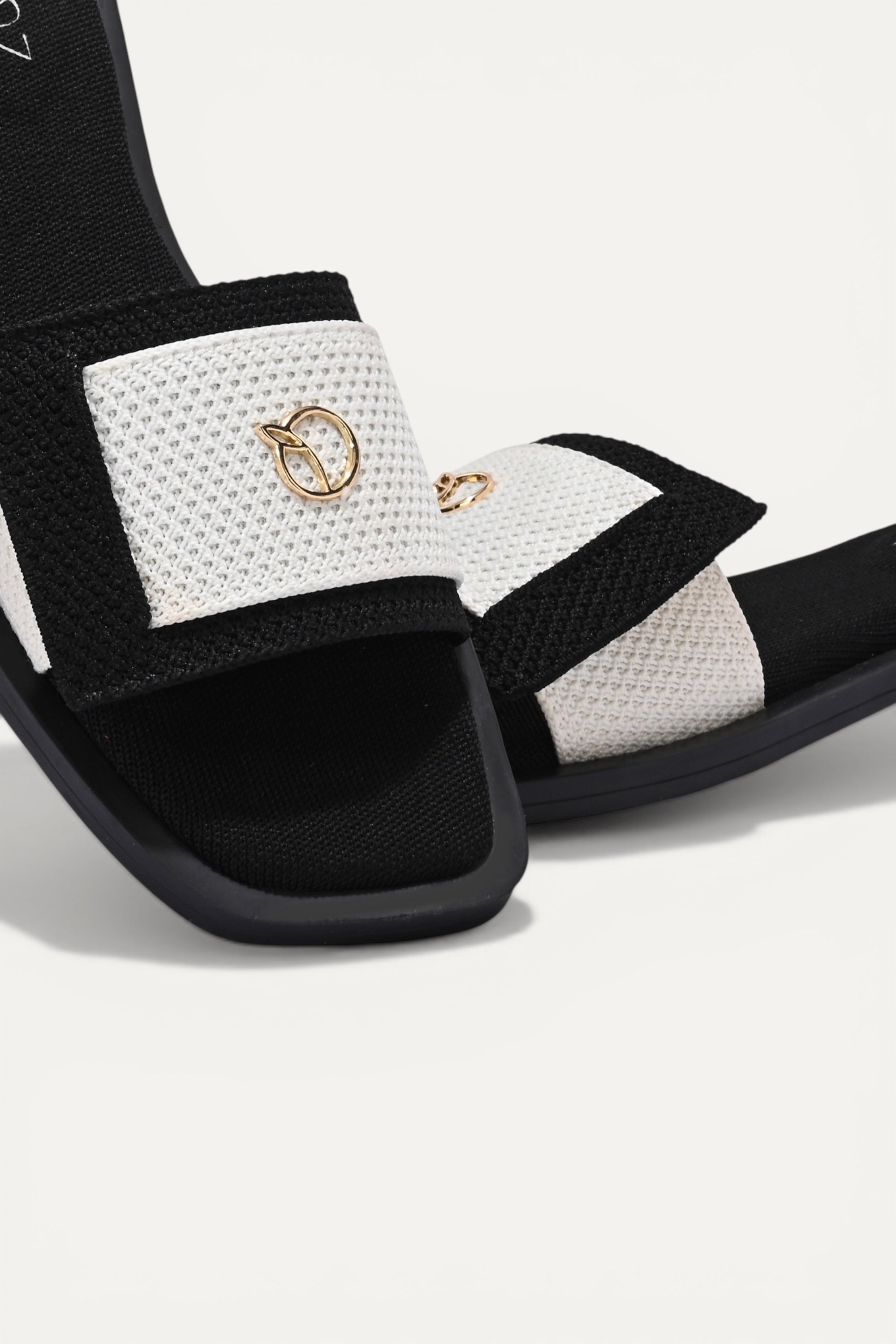 Rioz black and white knit origami slides with gold emblem detail. Asymmetrical fold design for a comfortable, breathable fit.